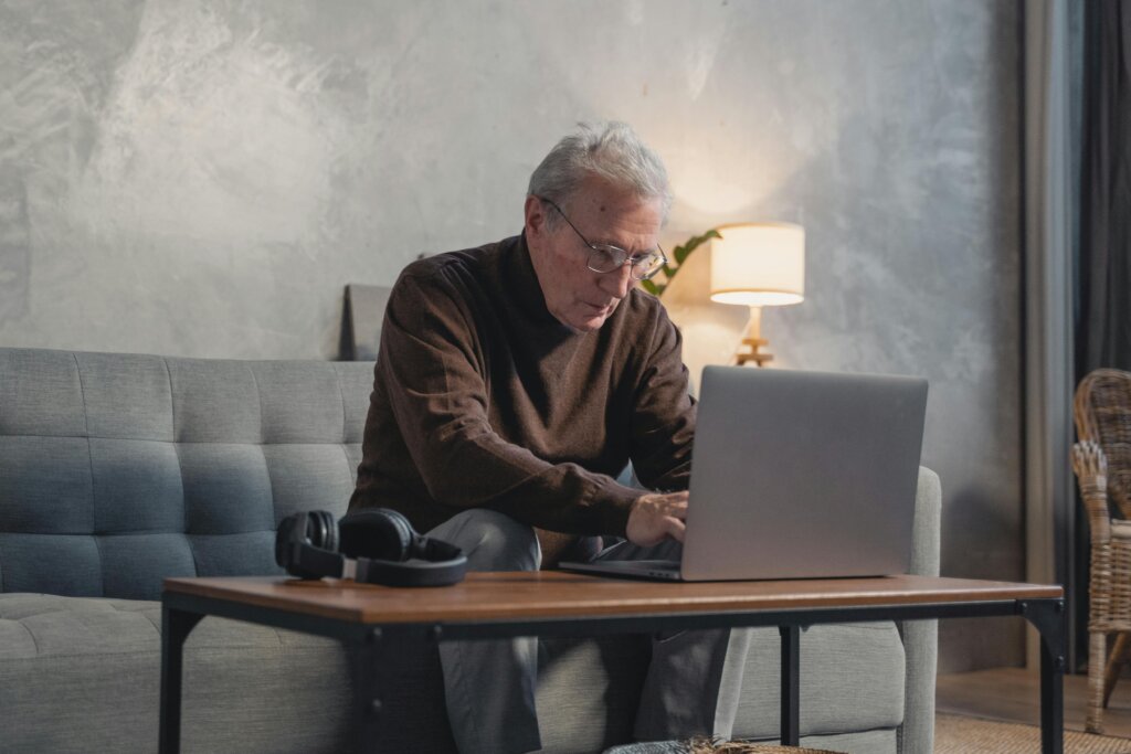 Ældre mand sidder på en sofa og arbejder på en bærbar computer med stort skærmbillede og tydelige skrifttyper for tilgængelighed.