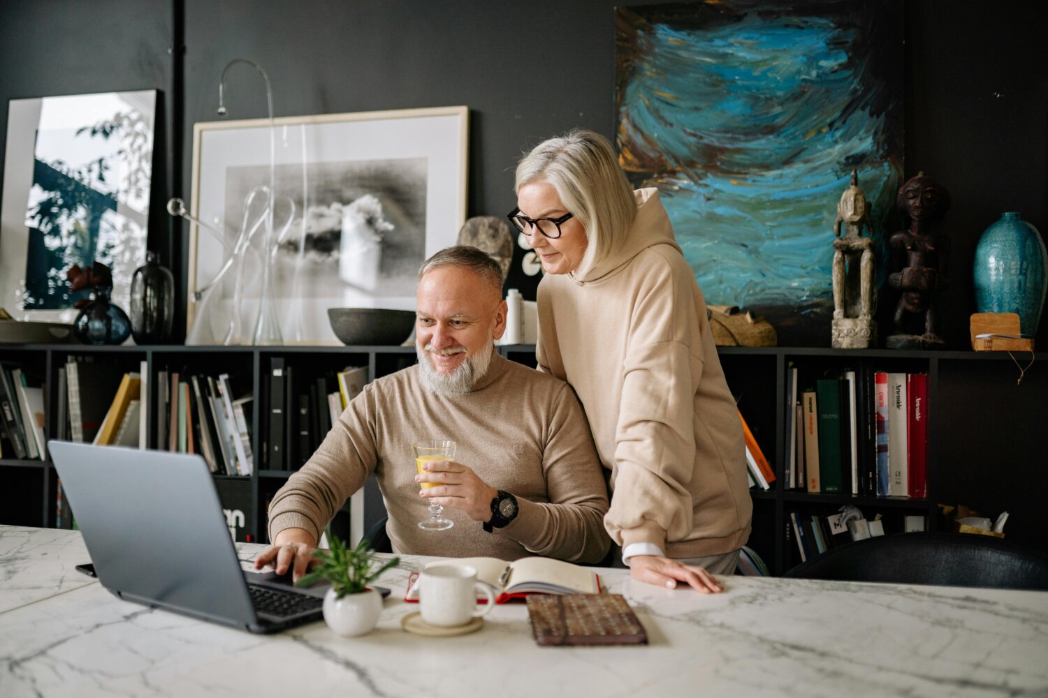 Ældre par smiler, mens de kigger på en bærbar computer ved et bord med bøger og kunstgenstande i baggrunden.