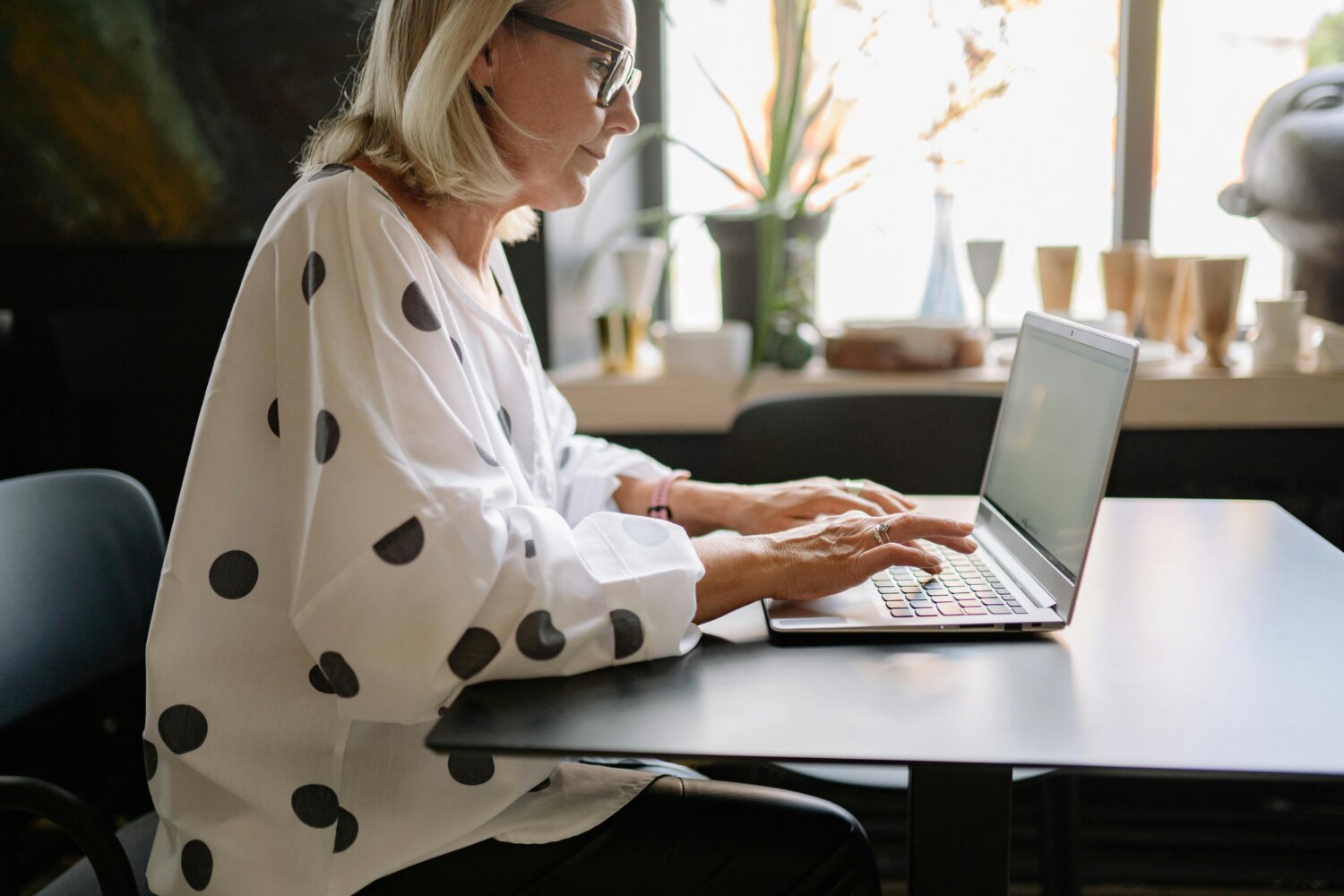 Kvinde med briller arbejder på en bærbar computer ved et bord i et lyst rum.