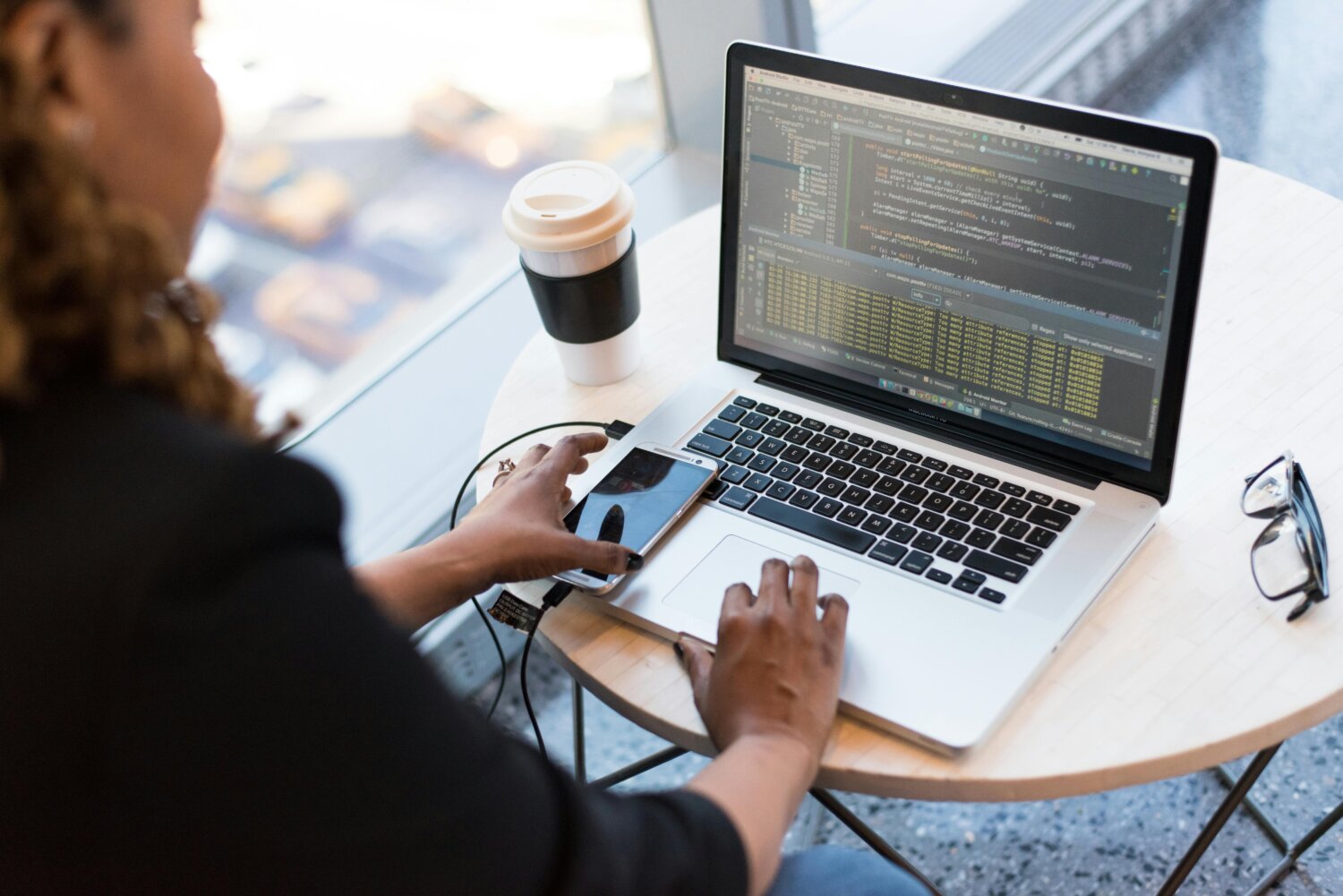 En person arbejder på en bærbar computer med koder på skærmen, har en smartphone tilsluttet, og en kaffekop står på bordet ved siden af.