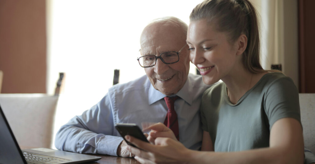 Ung kvinde og ældre mand sidder sammen ved et bord med en bærbar computer, mens de smiler og ser på en smartphone.