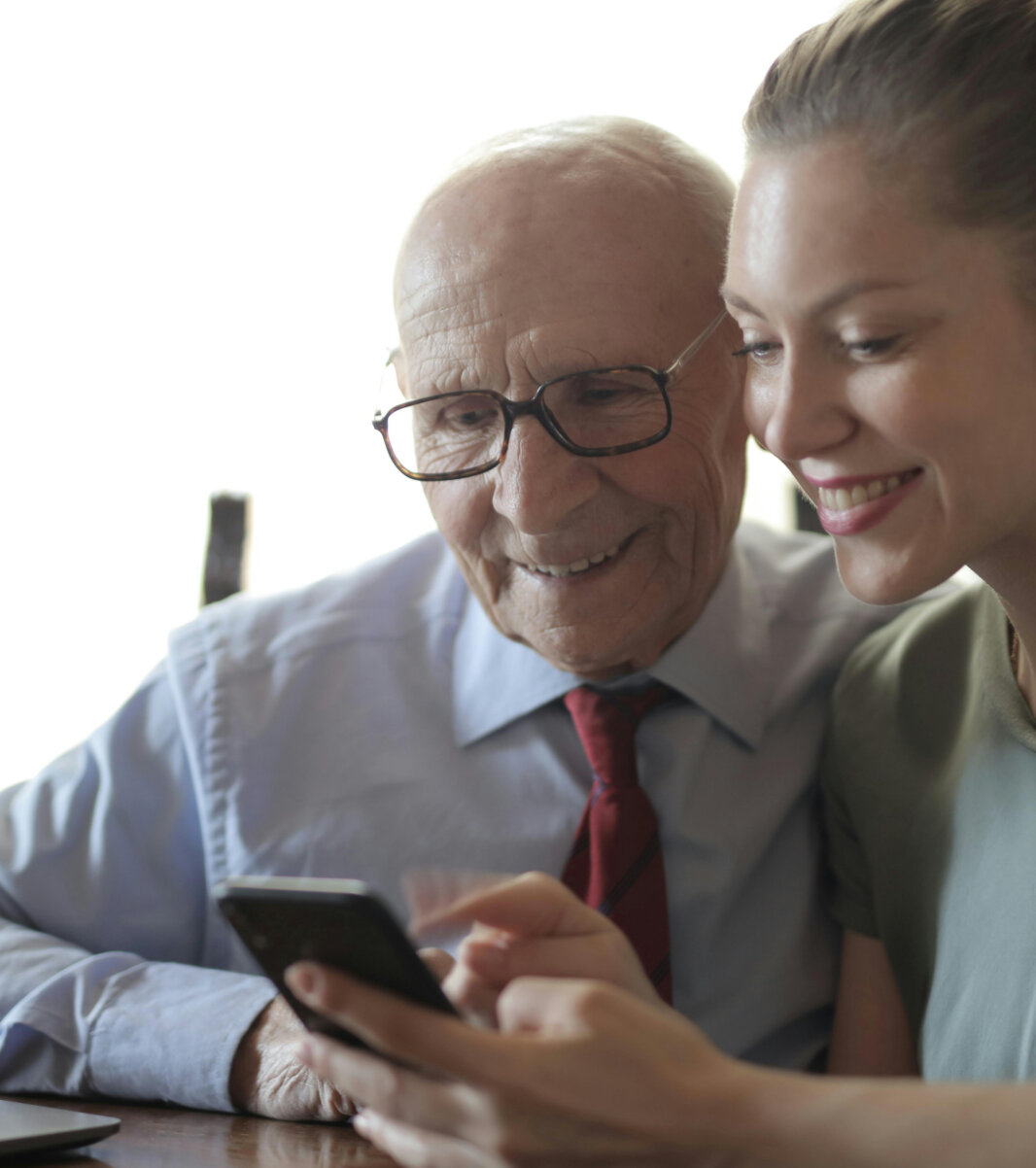 Ung kvinde og ældre mand sidder sammen ved et bord med en bærbar computer, mens de smiler og ser på en smartphone.