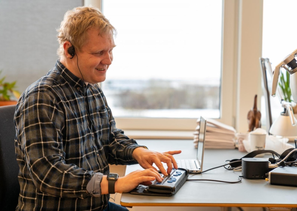 Mand med høretelefoner bruger skærmlæser på en bærbar computer på kontor, smiler og arbejder fokuseret.