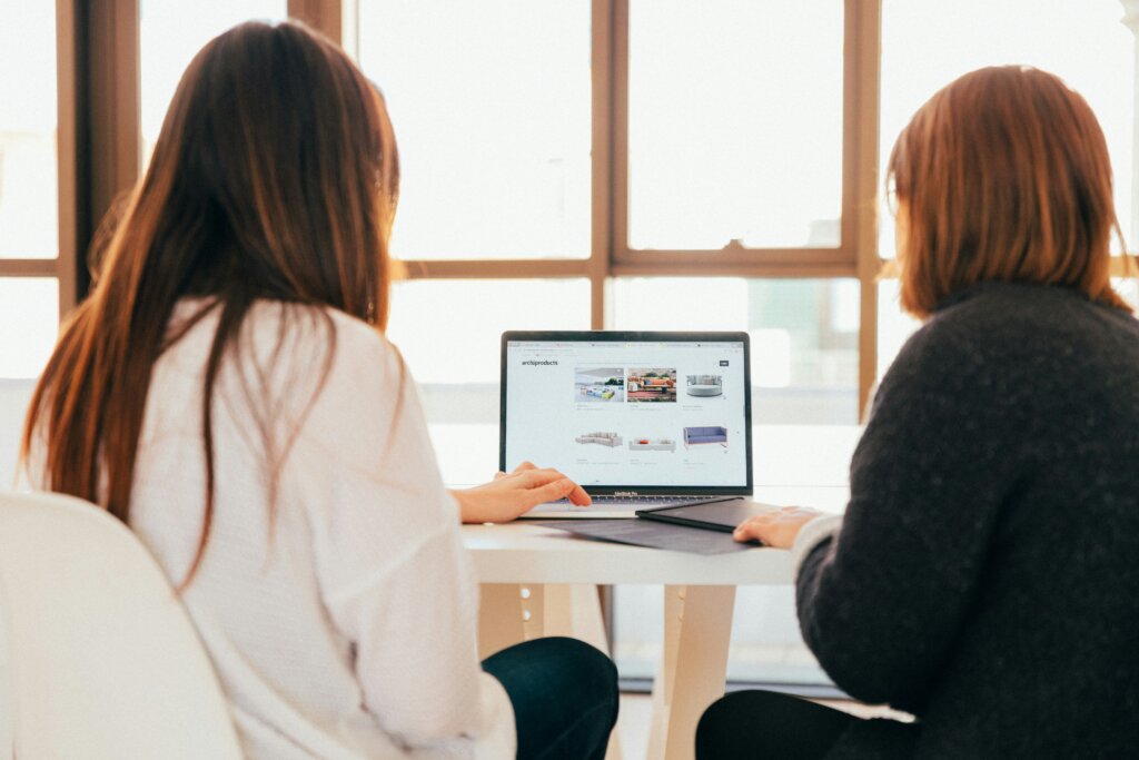 To personer sidder ved et bord og ser på en bærbar computer, der viser en webshop med møbler.