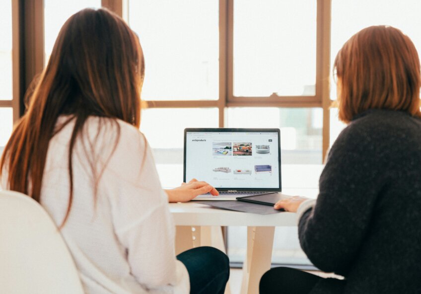 To personer sidder ved et bord og ser på en bærbar computer, der viser en webshop med møbler.