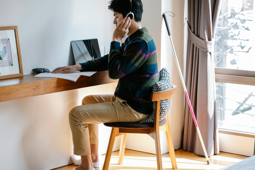 En mand sidder ved et skrivebord og arbejder med høretelefoner på. Han har en stok stående ved siden af sig, hvilket indikerer, at han er blind eller svagtseende. Der er en tablet på skrivebordet, og han hviler hånden på et dokument.