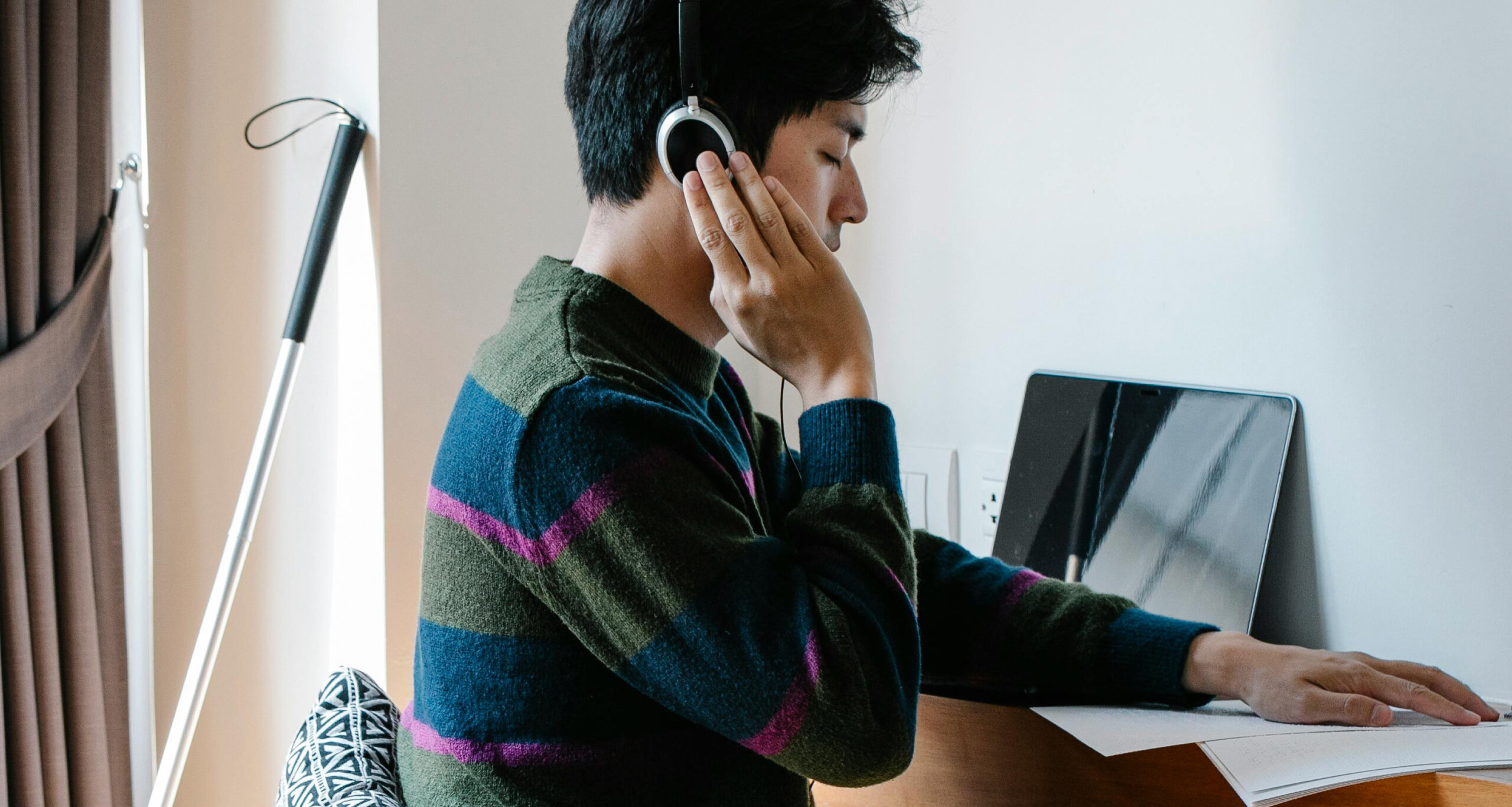 En mand med høretelefoner sidder ved et skrivebord og arbejder på en bærbar computer, med en stok til blindestøtte stående ved siden af.