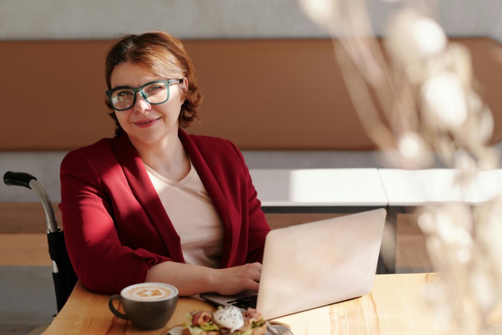 Kvinde i kørestol smiler, mens hun arbejder på en bærbar computer ved et bord i en café med en kop kaffe foran sig.