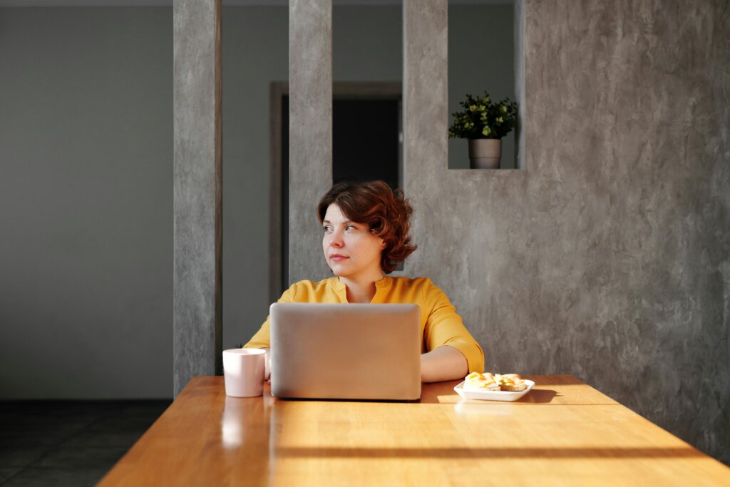 En kvinde i en gul bluse sidder ved et træbord med en bærbar computer foran sig. Hun kigger til siden og har en kaffekop og en tallerken med småkager ved siden af sig. Baggrunden består af en grå væg med en plante i en niche. Sollys skinner ind gennem et vindue og lyser scenen op.