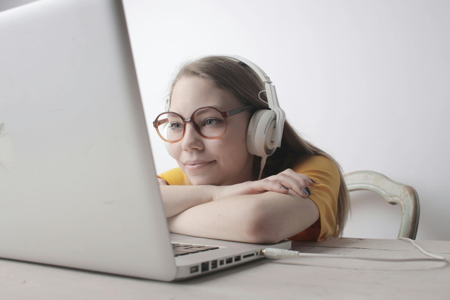 En ung kvinde med briller og hovedtelefoner smiler, mens hun kigger på sin bærbare computer. Hun hviler hovedet på armene ved et bord.