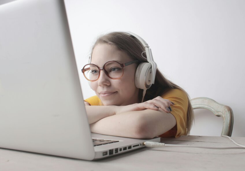 En ung kvinde med briller og hovedtelefoner smiler, mens hun kigger på sin bærbare computer. Hun hviler hovedet på armene ved et bord.