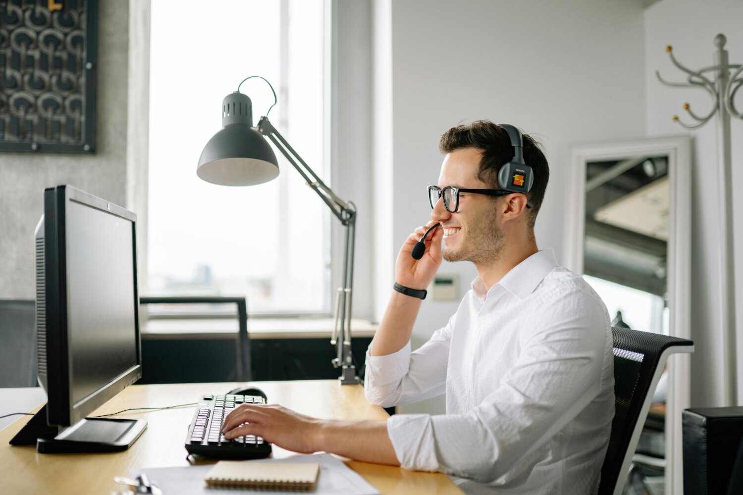 En smilende mand med briller og headset sidder ved et skrivebord og arbejder på en computer. Han taler i mikrofonen og kigger på skærmen.