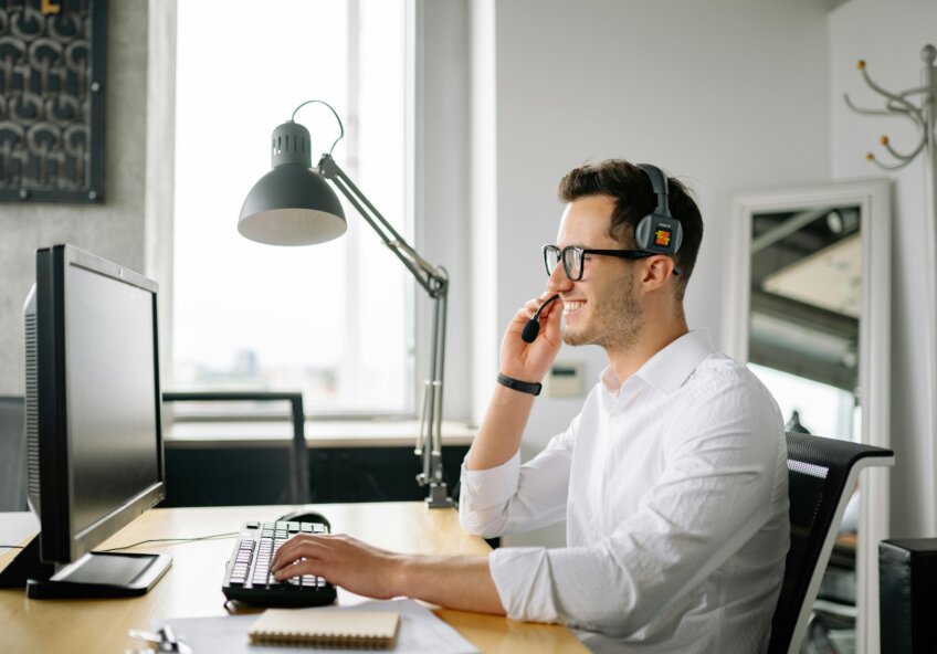 En smilende mand med briller og headset sidder ved et skrivebord og arbejder på en computer. Han taler i mikrofonen og kigger på skærmen.