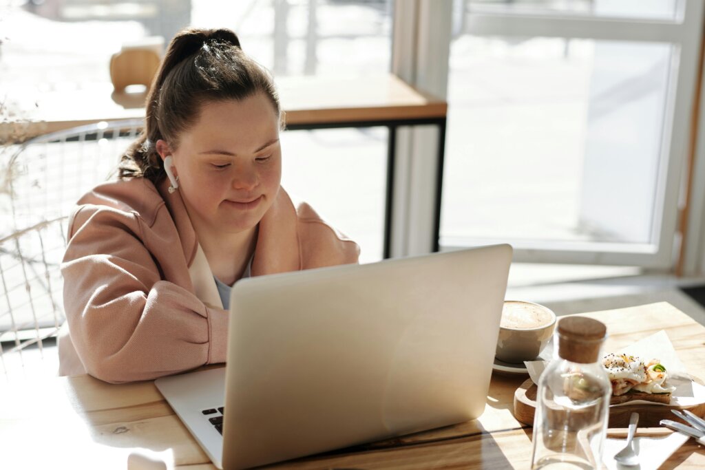 En ung kvinde med Downs syndrom arbejder på en bærbar computer i en lys café, med en kop kaffe og en sandwich på bordet.