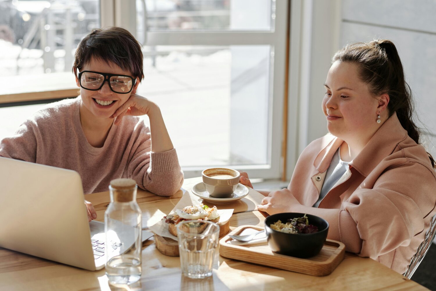 To kvinder sidder ved et cafébord med bærbar computer og mad foran sig, smilende og engagerede i samtale. Sollys strømmer ind gennem vinduet.