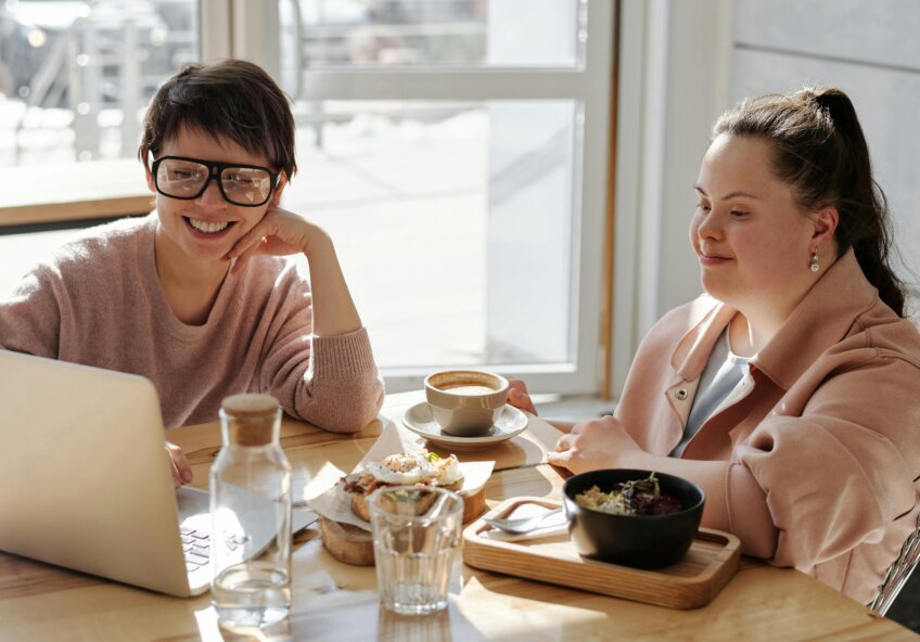 To kvinder sidder ved et cafébord med bærbar computer og mad foran sig, smilende og engagerede i samtale. Sollys strømmer ind gennem vinduet.