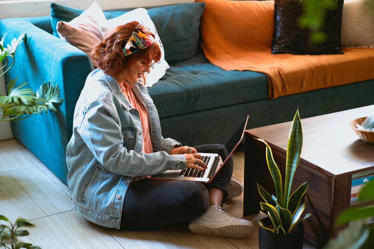 En kvinde med farverig hovedbeklædning sidder på gulvet foran en sofa og arbejder på en bærbar computer ved et lavt bord omgivet af stueplanter.