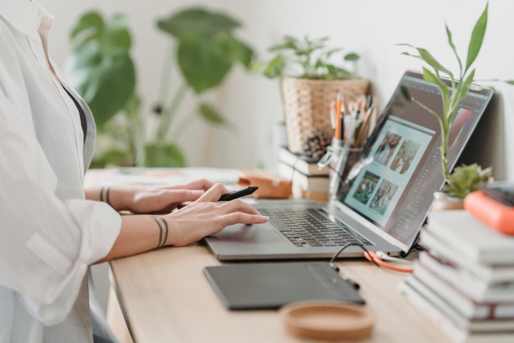 En person arbejder ved en bærbar computer på et skrivebord fyldt med planter, bøger og skriveredskaber. 