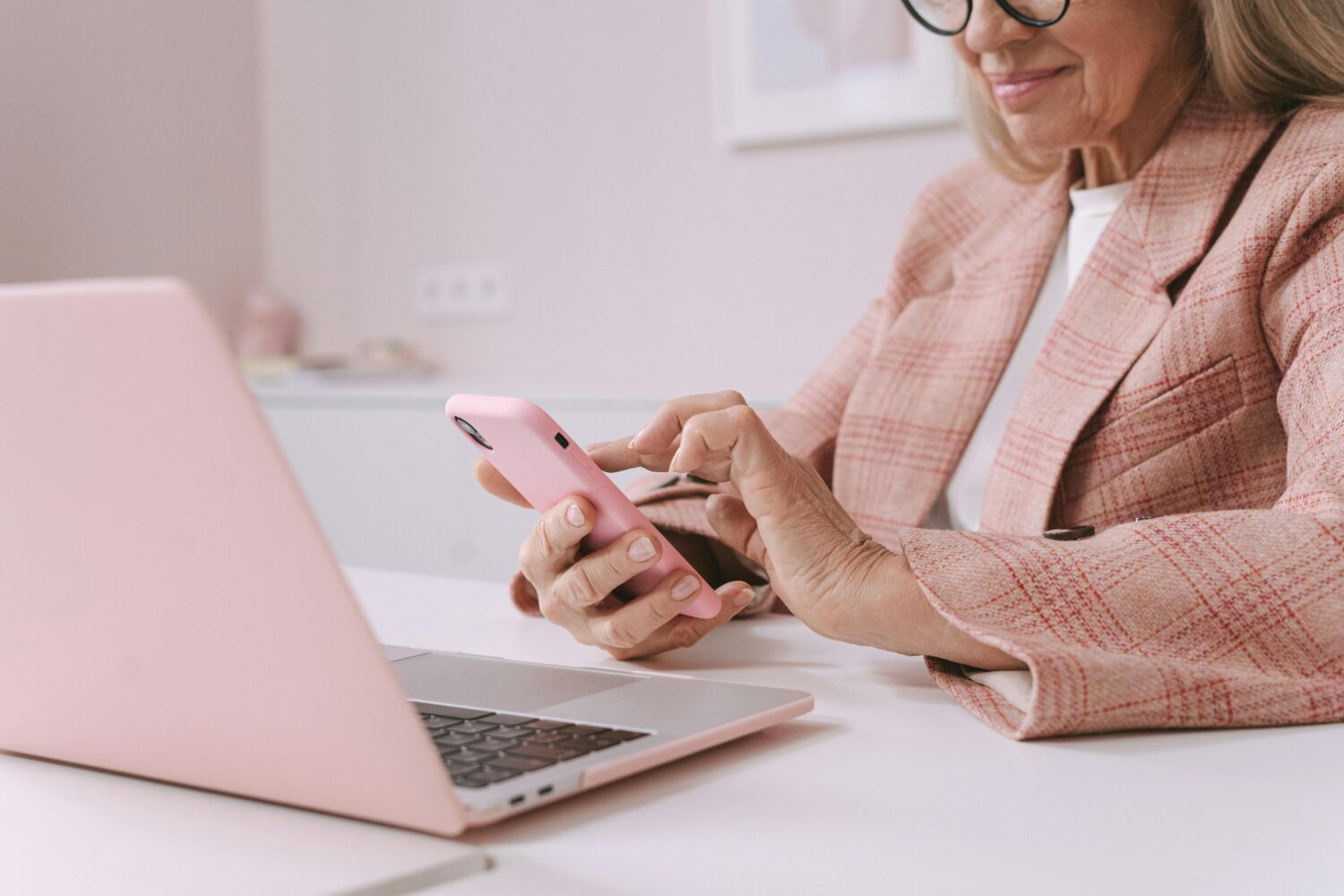 Ældre kvinde i ternet jakke arbejder på en bærbar computer og bruger en smartphone, begge i pink, ved et skrivebord.