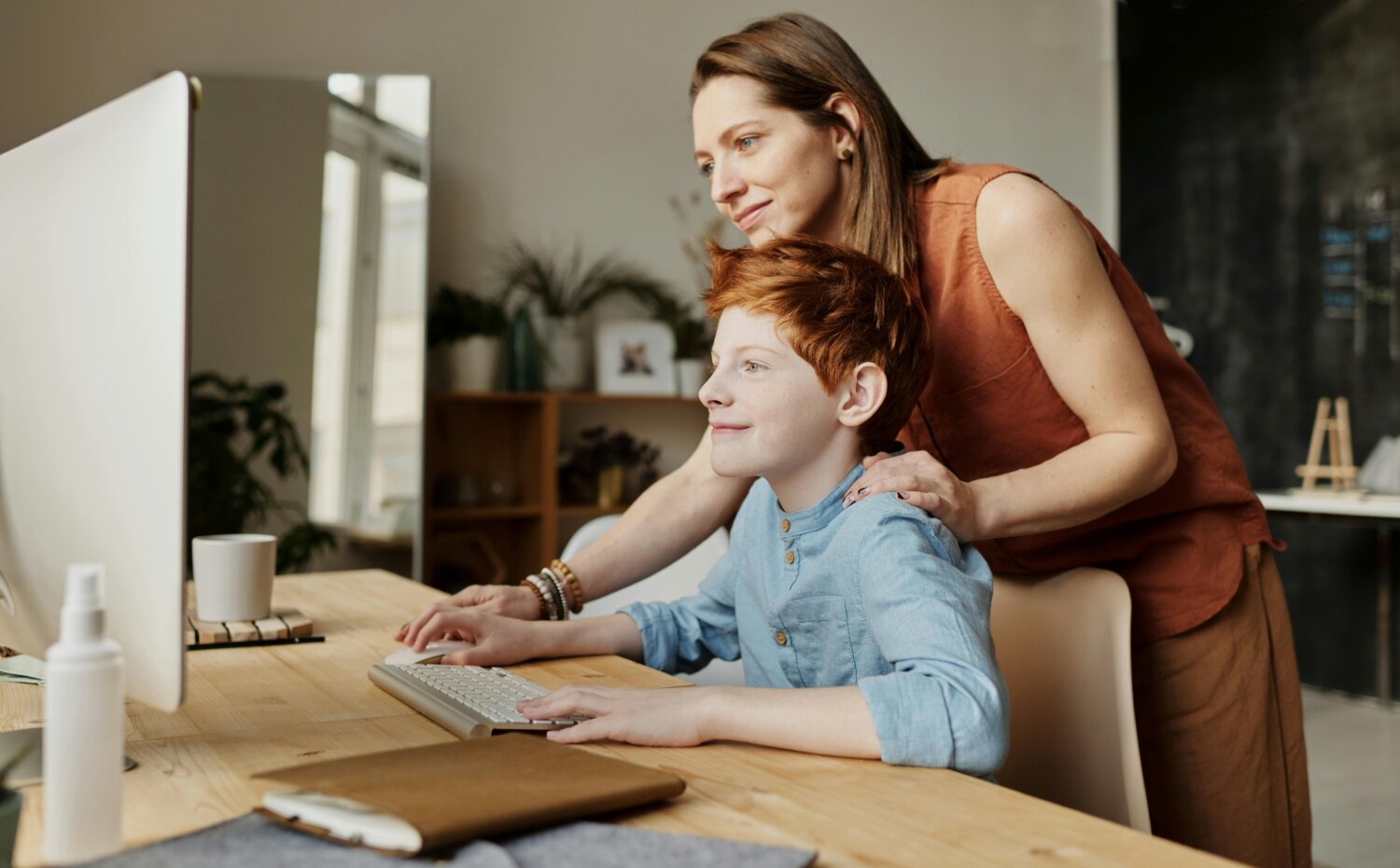 En kvinde står bag en dreng, der sidder ved et skrivebord og arbejder på en computer, mens hun smilende kigger med.