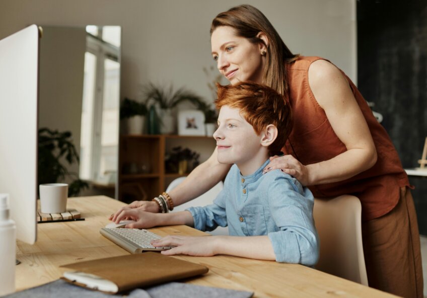 En kvinde står bag en dreng, der sidder ved et skrivebord og arbejder på en computer, mens hun smilende kigger med.