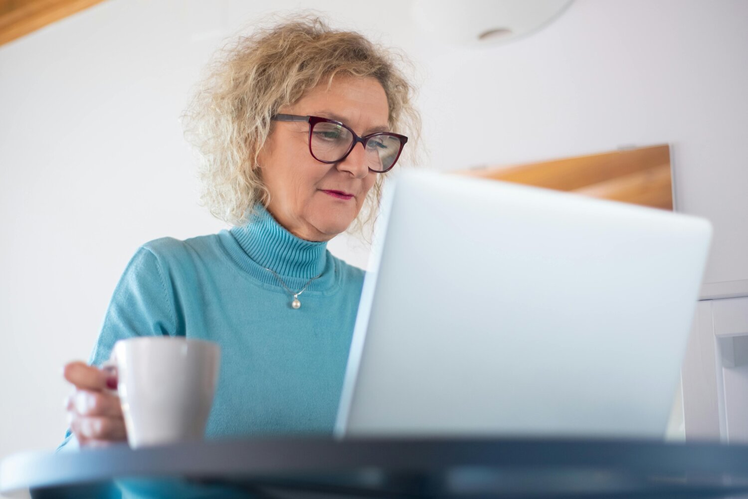 Ældre kvinde med briller arbejder på en bærbar computer og holder en kop kaffe.