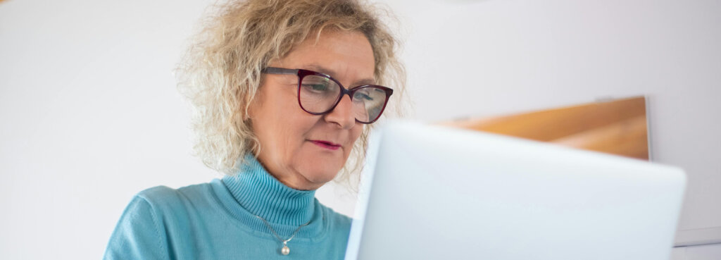 Ældre kvinde med briller arbejder på en bærbar computer og holder en kop kaffe.