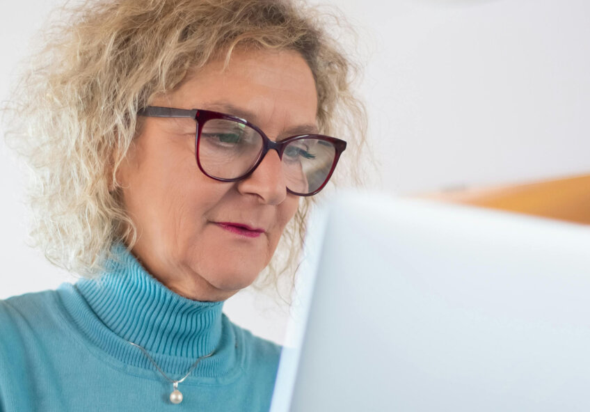 Ældre kvinde med briller arbejder på en bærbar computer og holder en kop kaffe.