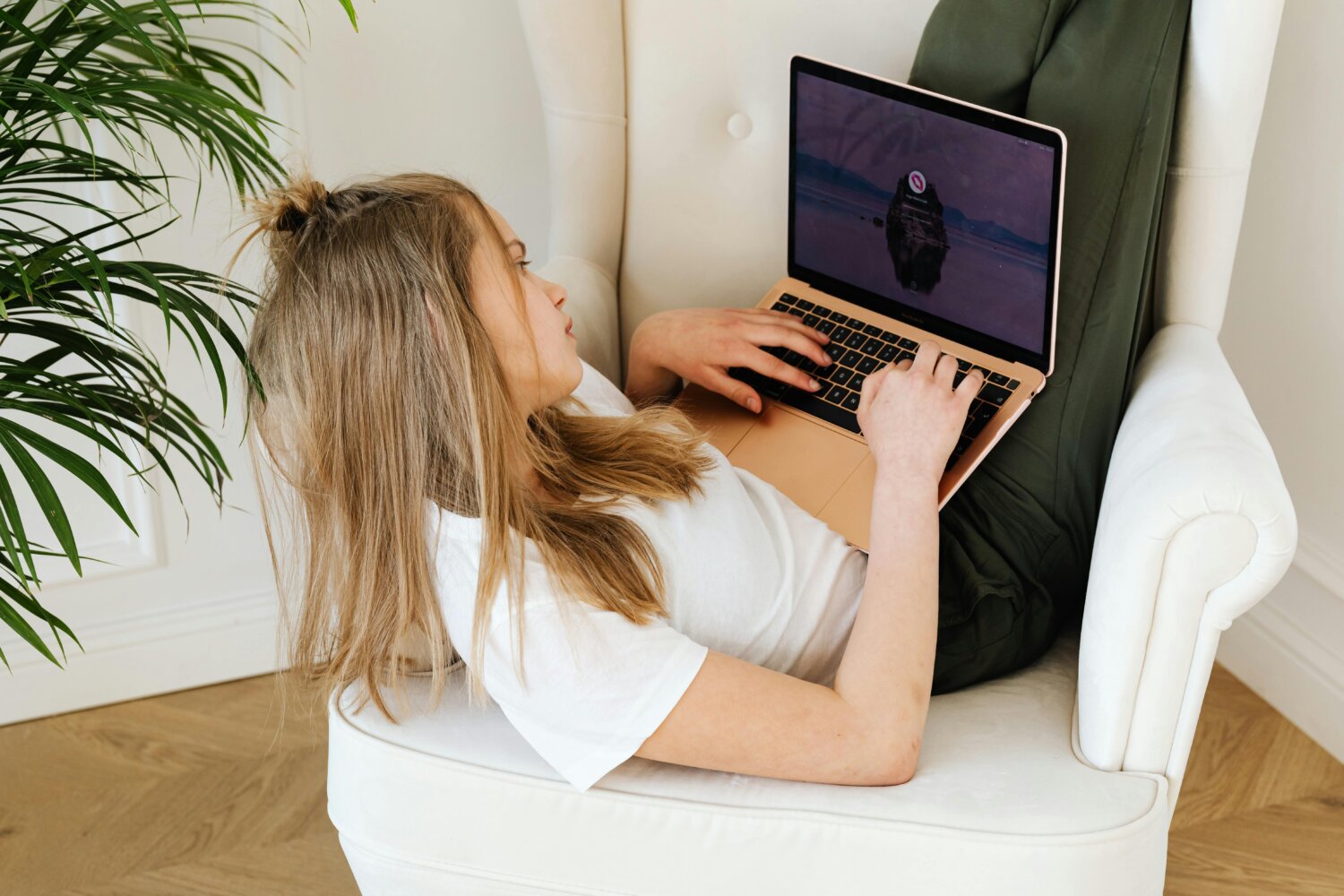 En ung pige ligger afslappet i en stol med benene oppe, mens hun arbejder på en bærbar computer, omgivet af en grøn plante.