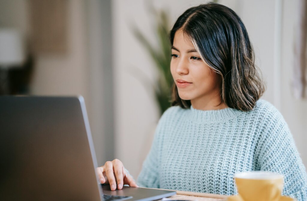 Kvinde i en lyseblå sweater arbejder koncentreret på en bærbar computer med en gul kop i forgrunden.