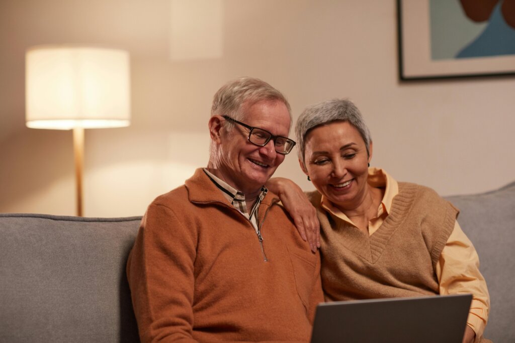 Ældre par sidder sammen på en sofa og smiler, mens de ser på en bærbar computer.