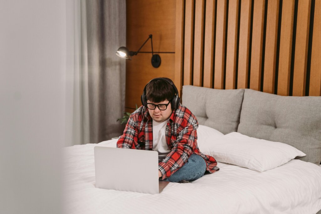 En person med hovedtelefoner og briller sidder på en seng og arbejder på en bærbar computer.