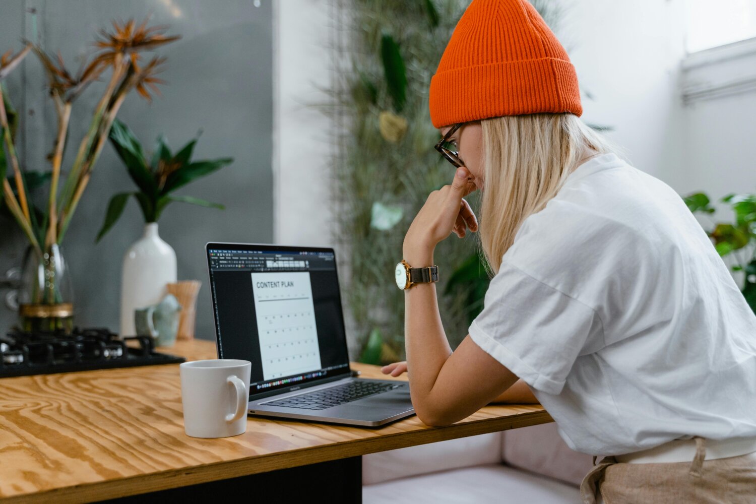 En kvinde med langt blondt hår og en orange hue arbejder ved en bærbar computer. Hun har en hvid t-shirt på og sidder ved et træbord med en kop kaffe.