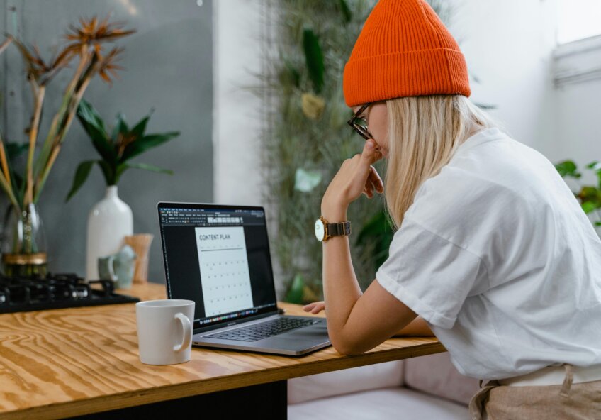 En kvinde med langt blondt hår og en orange hue arbejder ved en bærbar computer. Hun har en hvid t-shirt på og sidder ved et træbord med en kop kaffe.