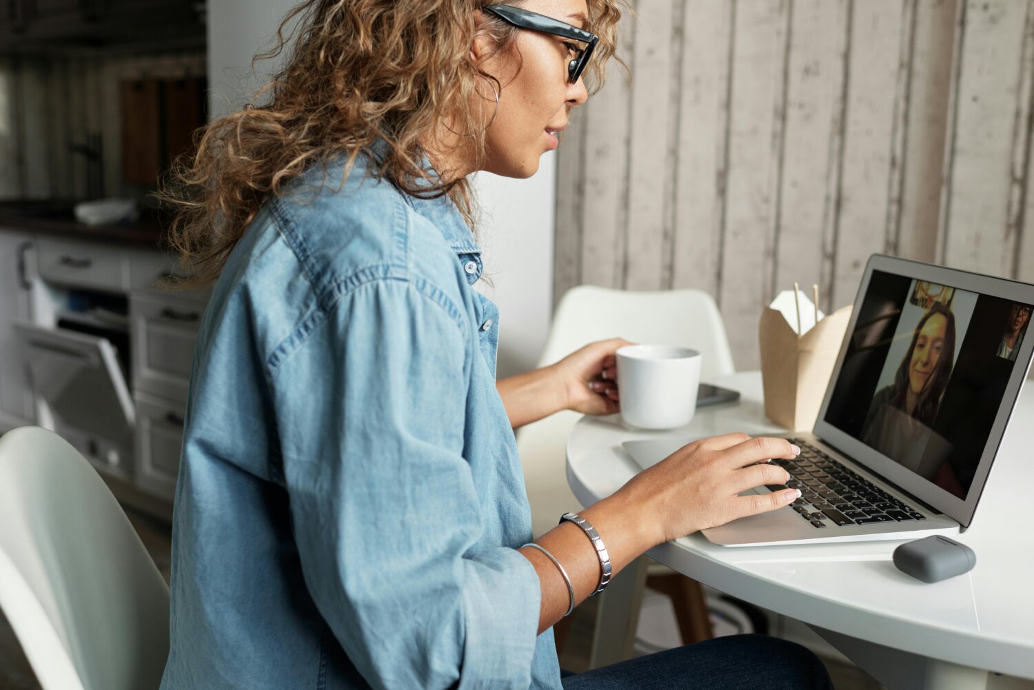 Kvinde med briller sidder ved et bord og deltager i et videomøde på sin bærbare computer, mens hun holder en kop kaffe.