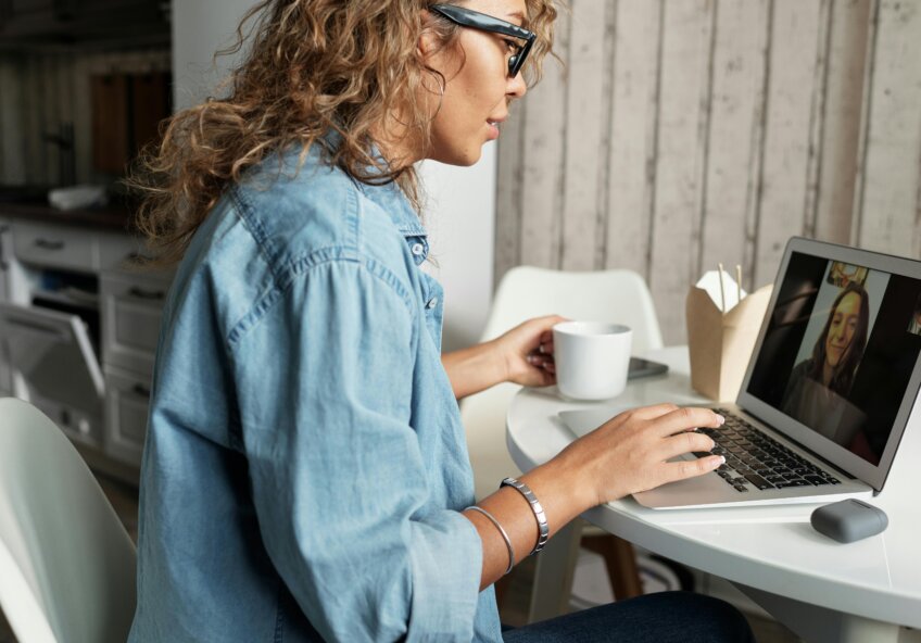 Kvinde med briller sidder ved et bord og deltager i et videomøde på sin bærbare computer, mens hun holder en kop kaffe.