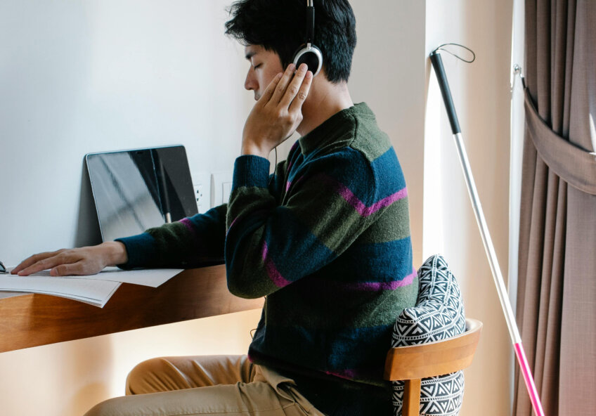 En mand med høretelefoner sidder ved et skrivebord og arbejder på en bærbar computer, med en stok til blindestøtte stående ved siden af.