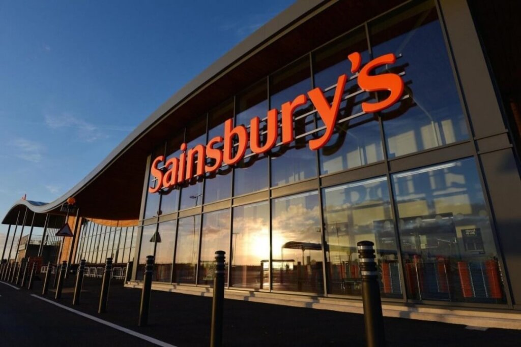 Billedet viser en moderne Sainsbury's butik med store glasvinduer, hvor solens refleksion kan ses. Bygningens facade er domineret af det velkendte røde Sainsbury's logo.